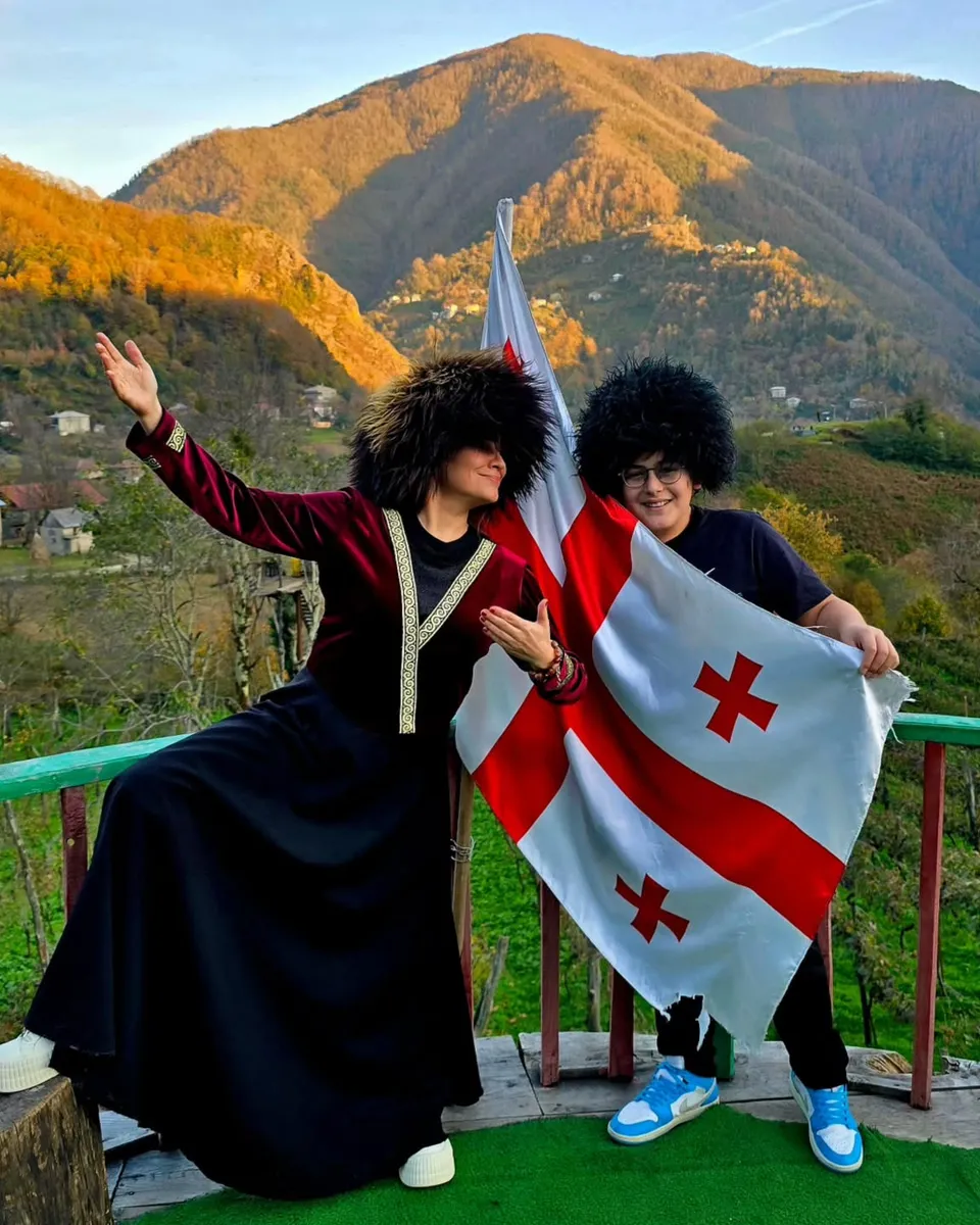 Two people in traditional papakha hats holding a Georgian flag on a wooden terrace, with autumn mountains and a village behind them